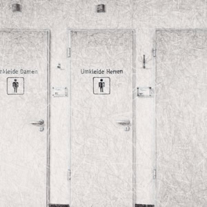 An artistic hand drawing by Herbert Stattler depicting the basement level of a tumor rehabilitation clinic. The scene shows a lone patient walking down a hallway lined with four doors, including marked locker rooms for men and women.